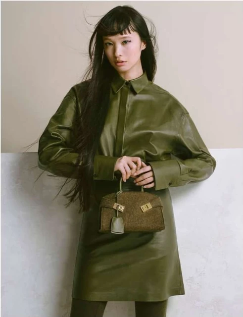 Asian model poses in an olive green, long-sleeved leather dress, holding a matching bag against a neutral two-tone background.