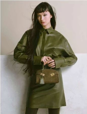 Asian model poses in an olive green, long-sleeved leather dress, holding a matching bag against a neutral two-tone background.