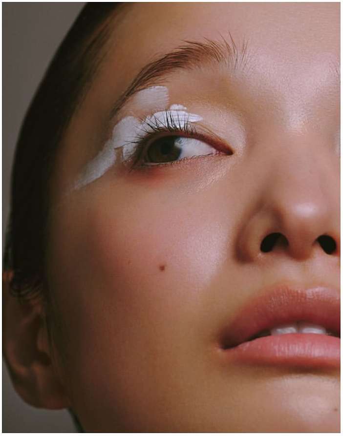 Close-up portrait of a woman with white paint accenting her upper eyelid and lashes, emphasizing her facial features and complexion.