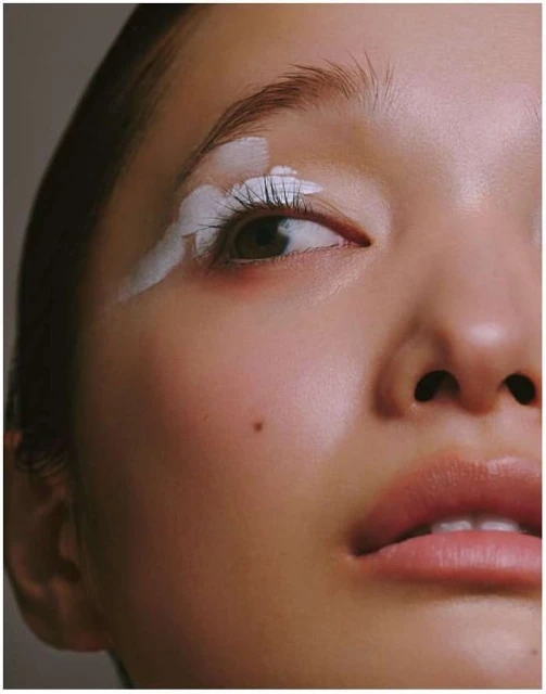 Close-up portrait of a woman with white paint accenting her upper eyelid and lashes, emphasizing her facial features and complexion.