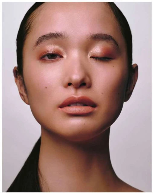 Close-up portrait of an Asian woman with fair skin and brown hair pulled back, wearing natural-toned makeup, and partially closing one eye.