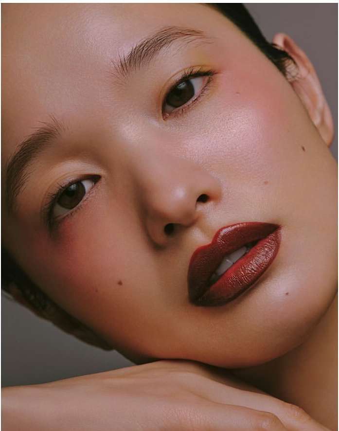 Close-up portrait of an Asian woman with dark red lipstick, glowing skin, and a mole on her face, resting her chin on her hand.