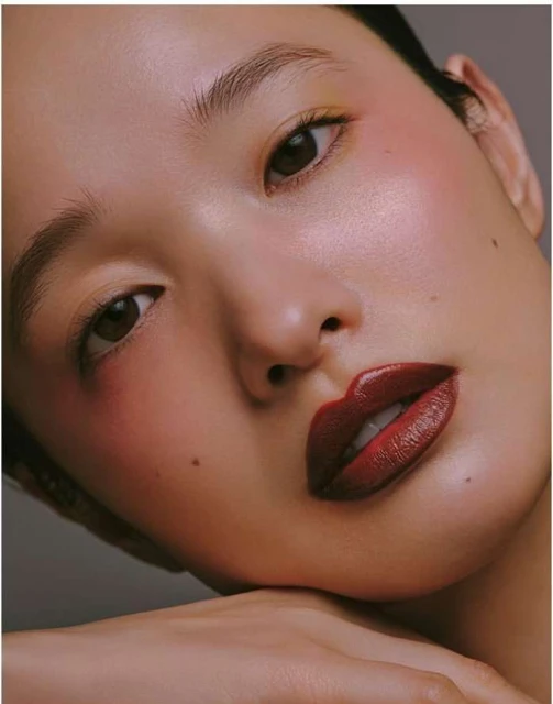 Close-up portrait of an Asian woman with dark red lipstick, glowing skin, and a mole on her face, resting her chin on her hand.