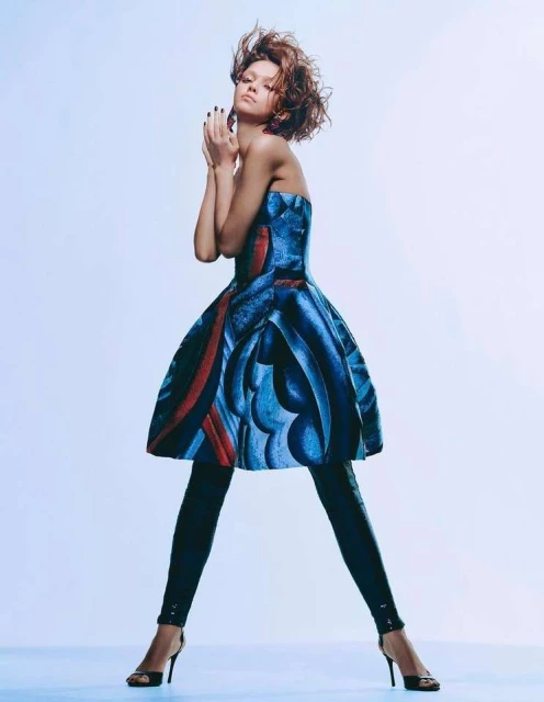 A woman in a blue patterned strapless dress with black leggings and high heels posing in a photo studio with a light blue backdrop.