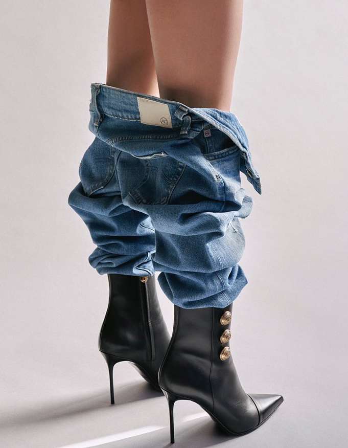 Woman wearing blue jeans and black heeled boots against a white background in a fashion editorial studio shot.