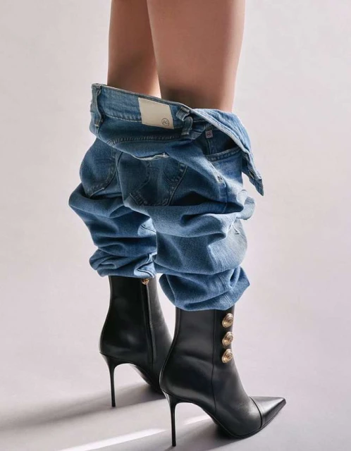 Woman wearing blue jeans and black heeled boots against a white background in a fashion editorial studio shot.