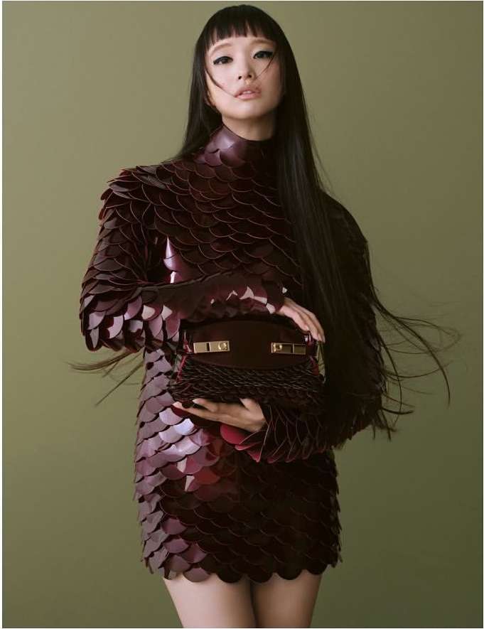 Woman in a burgundy, textured mini-dress with long, straight black hair, holding a brown handbag, against a solid green backdrop.