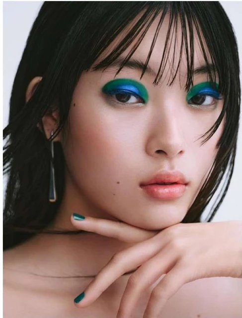 Close-up portrait of a young woman with dark hair, wearing blue and green eyeshadow, resting her chin on her manicured hand.
