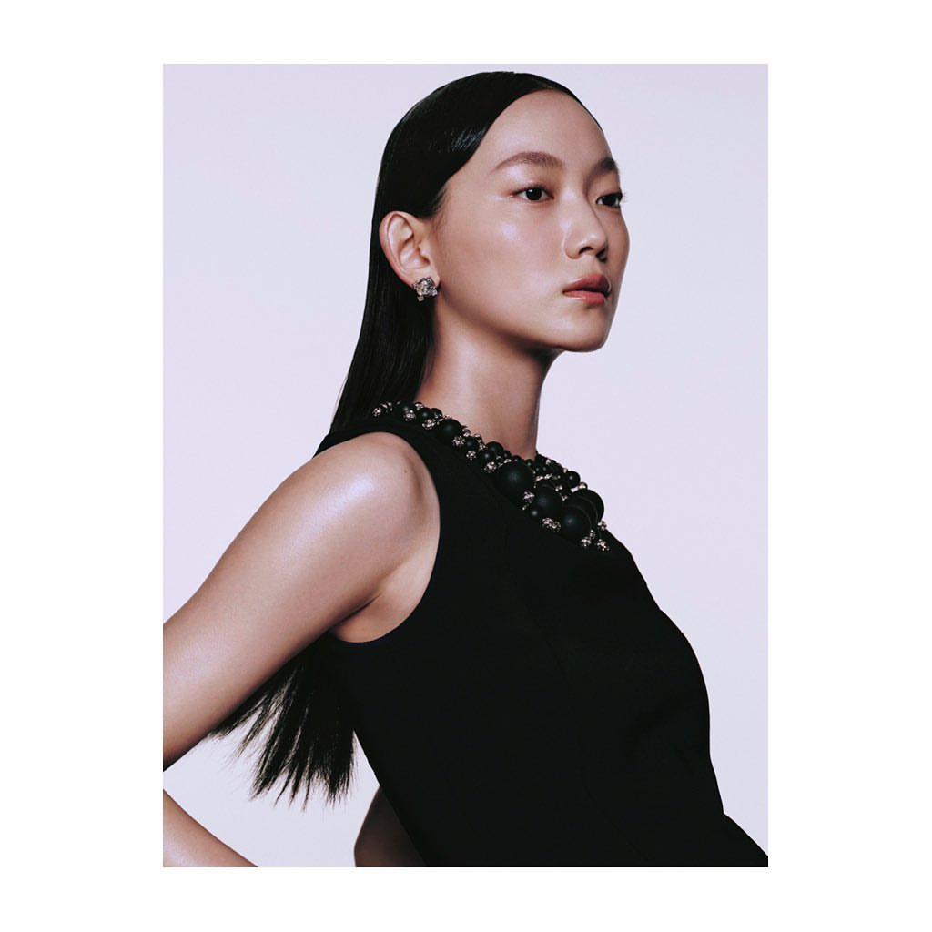 Portrait of an Asian woman with slicked-back hair wearing a black dress and statement jewelry in a photo studio setting with white background.
