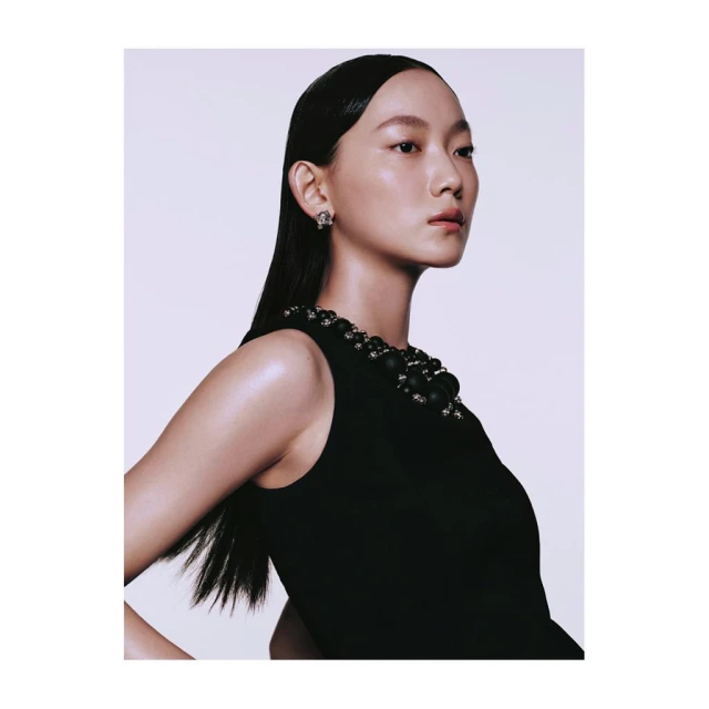 Portrait of an Asian woman with slicked-back hair wearing a black dress and statement jewelry in a photo studio setting with white background.