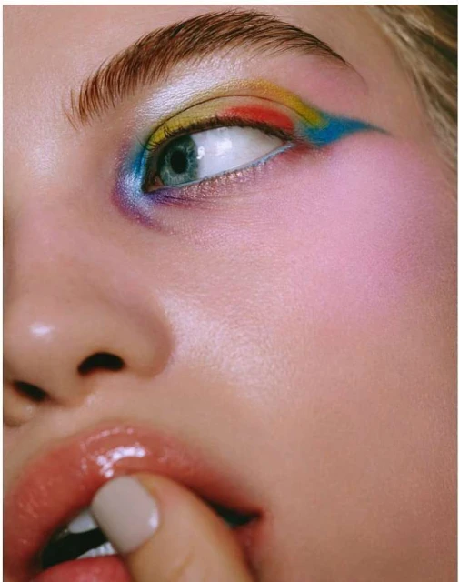 Close-up portrait of a young adult female with colorful eyeshadow, glossy lips, and a finger near her mouth in a beauty-focused shot.
