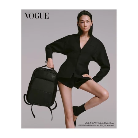 Fashion editorial shot of a woman in a black suit holding a Dell backpack against a grey background for a Vogue Japan advertisement.