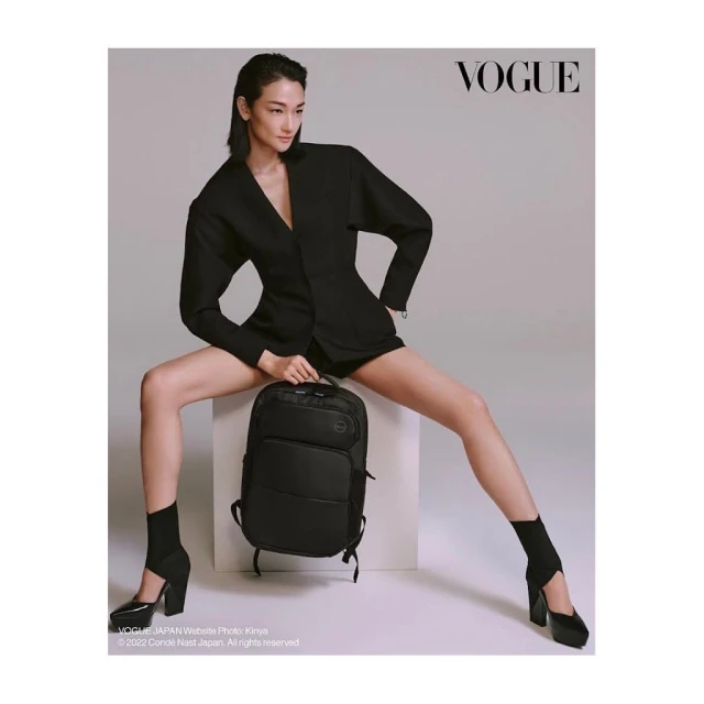 Asian female model poses with a black backpack while sitting on a cube in a studio with 'Vogue' logo at the top. 