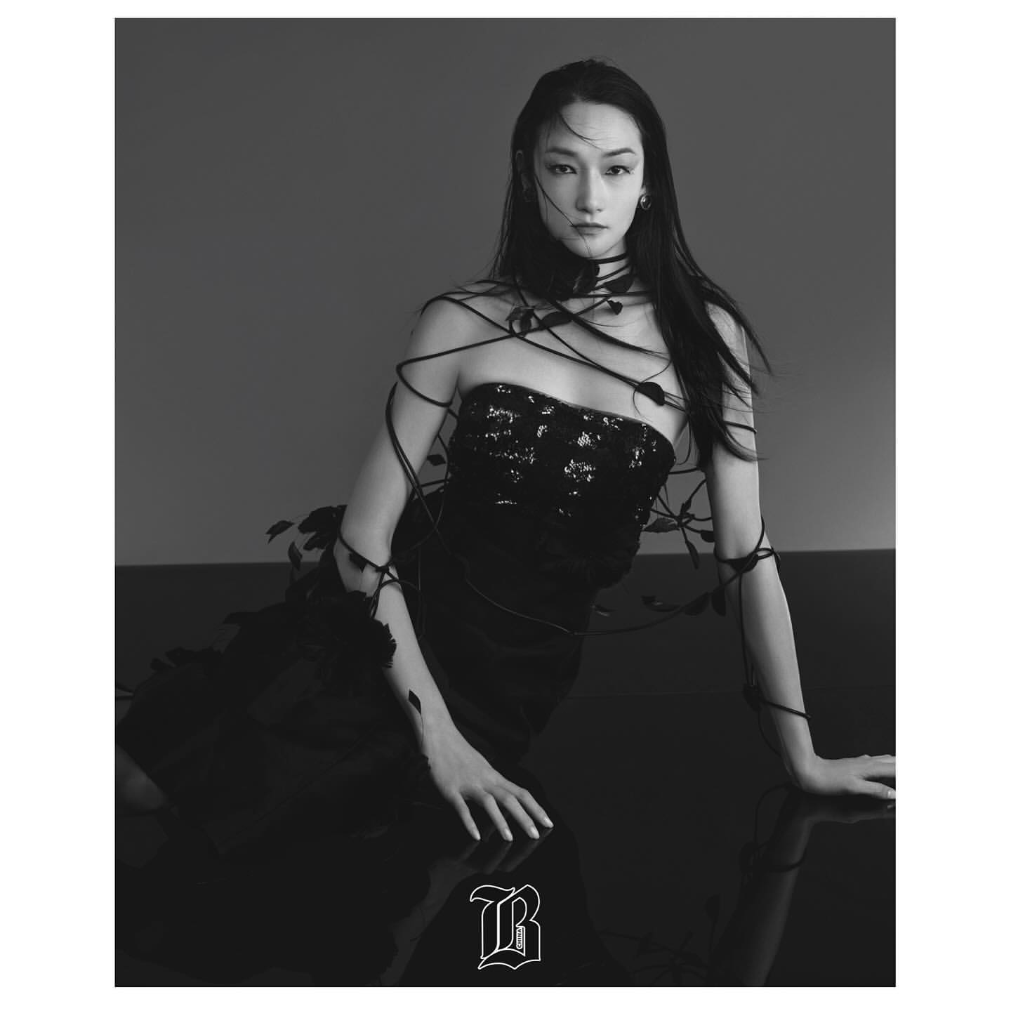 A fashionable young woman in a black dress and statement necklace poses indoors with long dark hair and dramatic studio lighting.