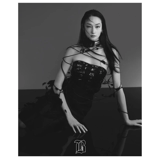 A fashionable young woman in a black dress and statement necklace poses indoors with long dark hair and dramatic studio lighting.