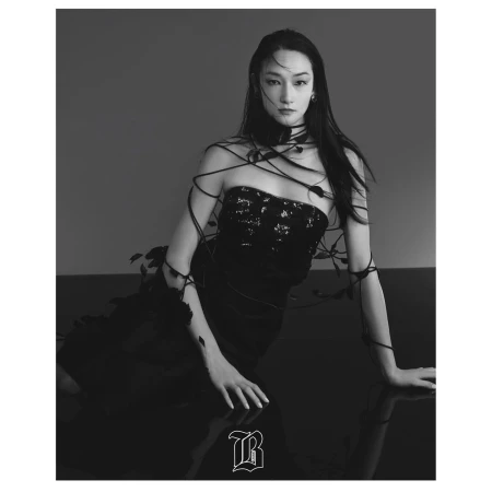 A fashionable young woman in a black dress and statement necklace poses indoors with long dark hair and dramatic studio lighting.