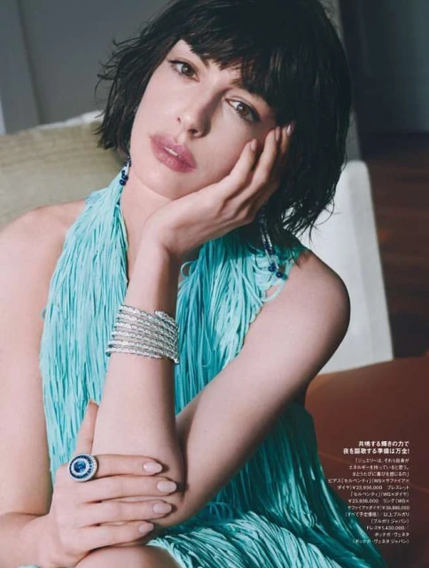 Anne Hathaway poses indoors wearing a fringed dress and jewelry, looking thoughtfully into the camera. Fashion and beauty editorial.