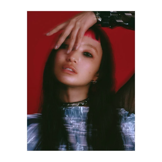 Eye-level portrait of a stylish young woman with pink bangs, wearing a studded choker and shimmering blue top against a red backdrop.