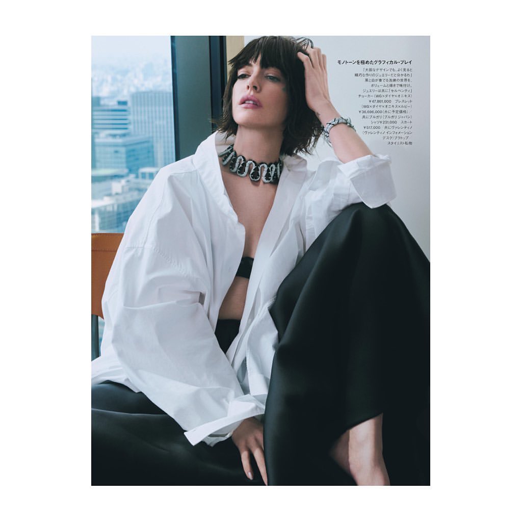 Fashion editorial shot of a brunette adult woman wearing a white shirt and jewelry, sitting against an urban background.