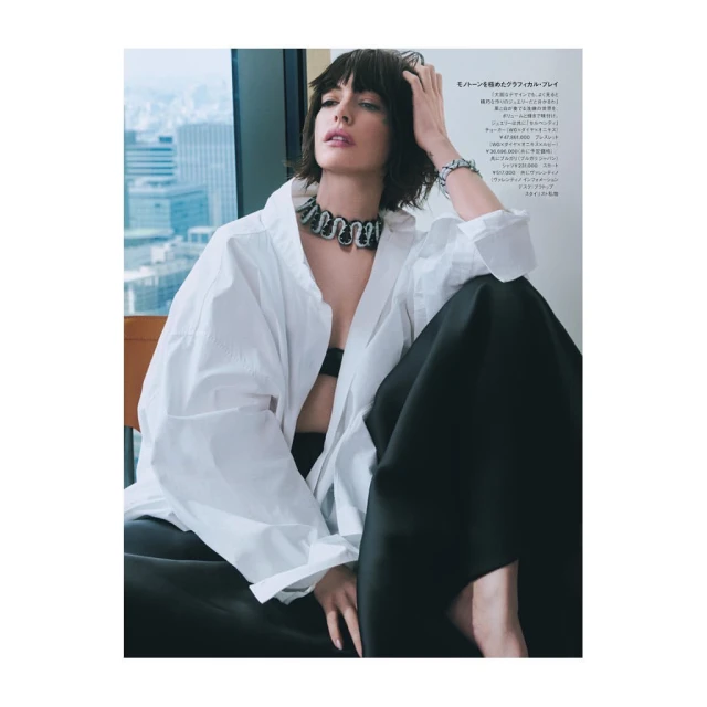 Fashion editorial shot of a brunette adult woman wearing a white shirt and jewelry, sitting against an urban background.
