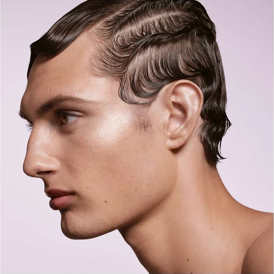 Close-up profile of a man with dark slicked-back hair styled in waves, side view, soft lighting and a clean, light backdrop.
