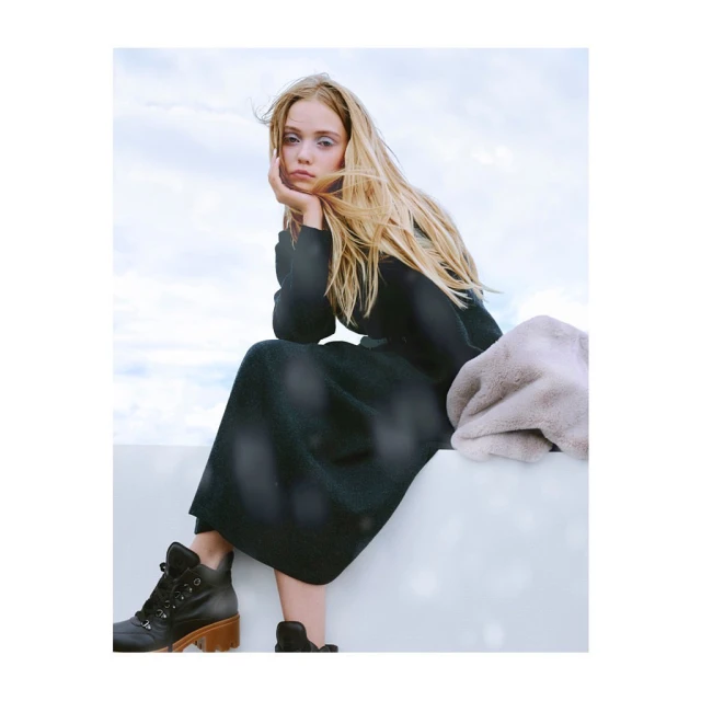 A blonde female fashion model sits outdoors wearing a long black dress and boots, with a light grey blanket and bright sky background.