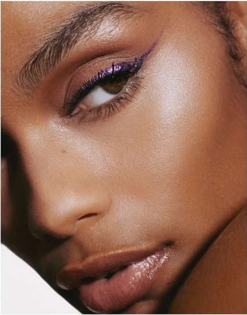 Close-up portrait of a woman with brown skin, brown eyes, purple eyeliner, and glossy lips, against a white background in a beauty shot.