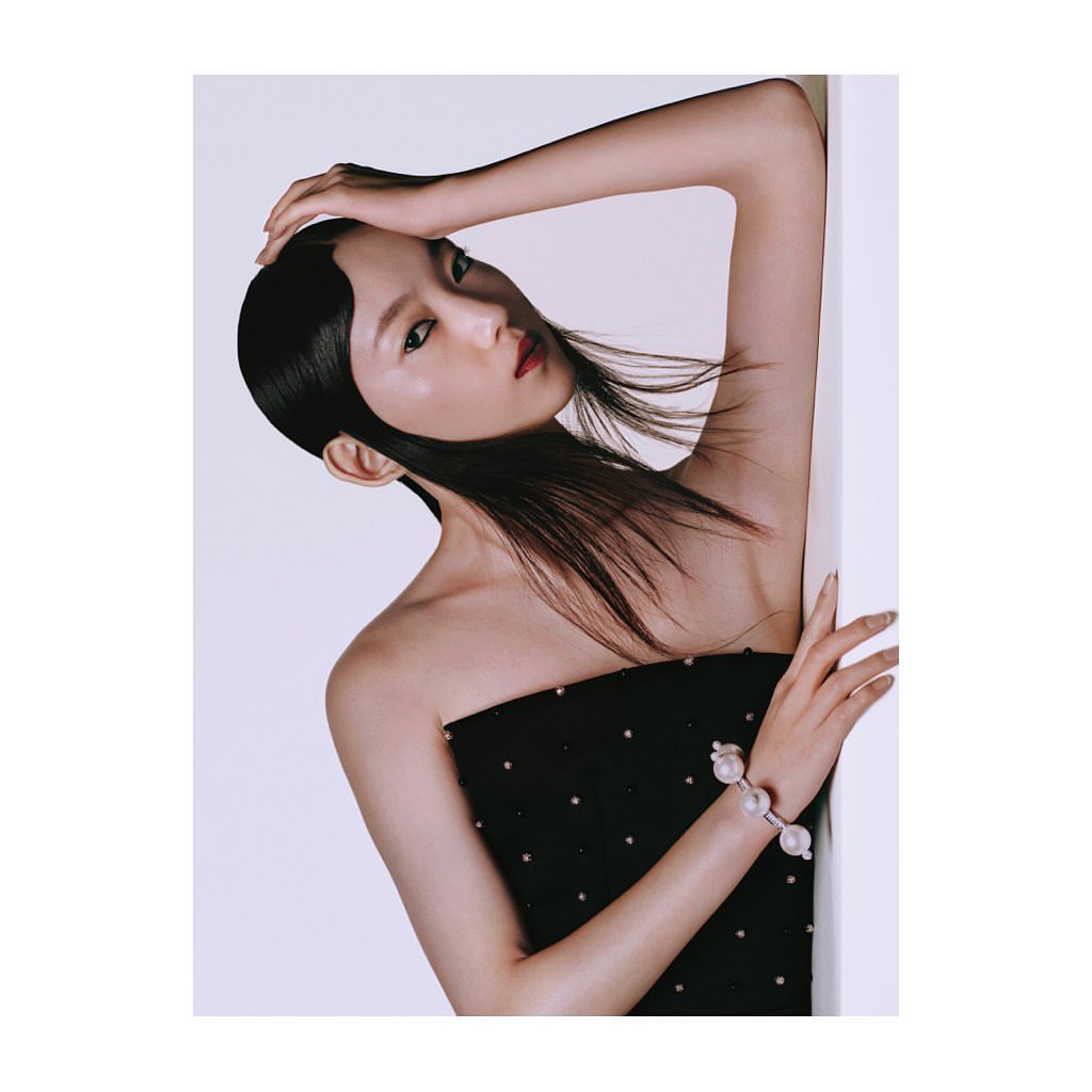 Elegant Asian woman poses in a strapless black dress with pearl jewelry against a clean, bright backdrop in a studio setting.