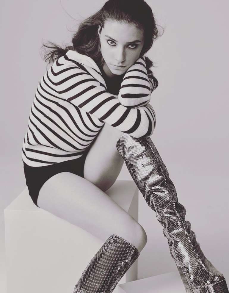 Striking black and white studio portrait of a woman in a striped sweater, black shorts, and high snakeskin boots, seated on a white cube.