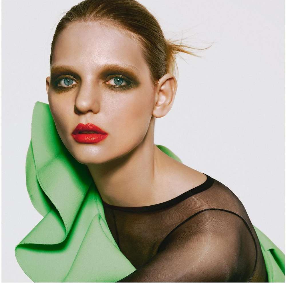 Close-up fashion shot of a fair-skinned woman with heavy makeup and a green top. She is facing forward against a white background.