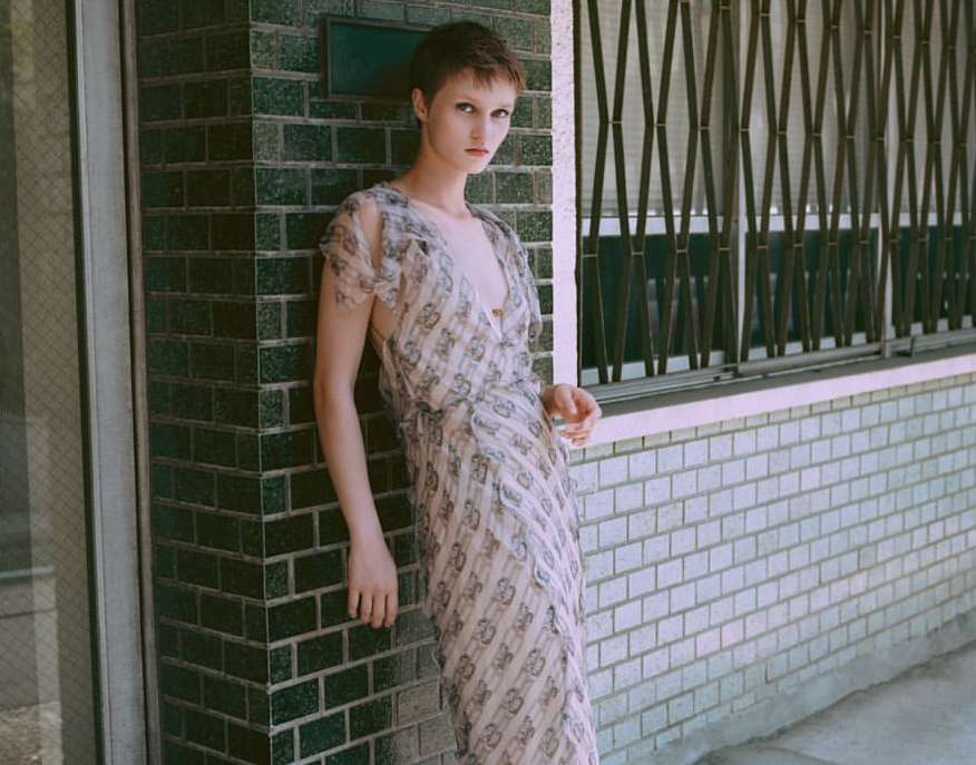 Woman in patterned dress poses against a brick wall with metal grate window in the background, neutral color grading, natural lighting.