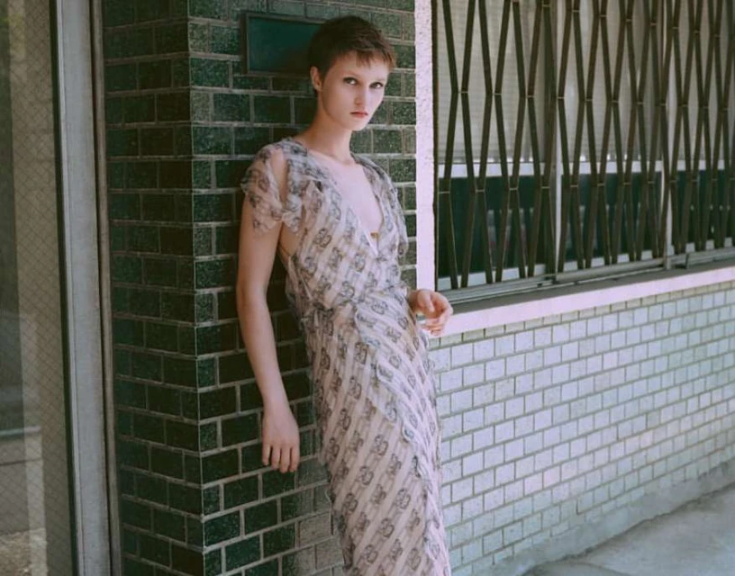 Woman in patterned dress poses against a brick wall with metal grate window in the background, neutral color grading, natural lighting.