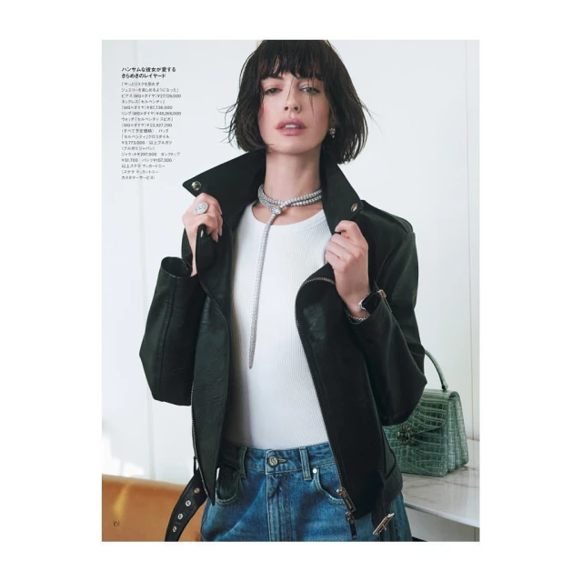 Anne Hathaway poses in a stylish editorial shot wearing a black leather jacket, white top, blue jeans, and diamond jewelry in studio.