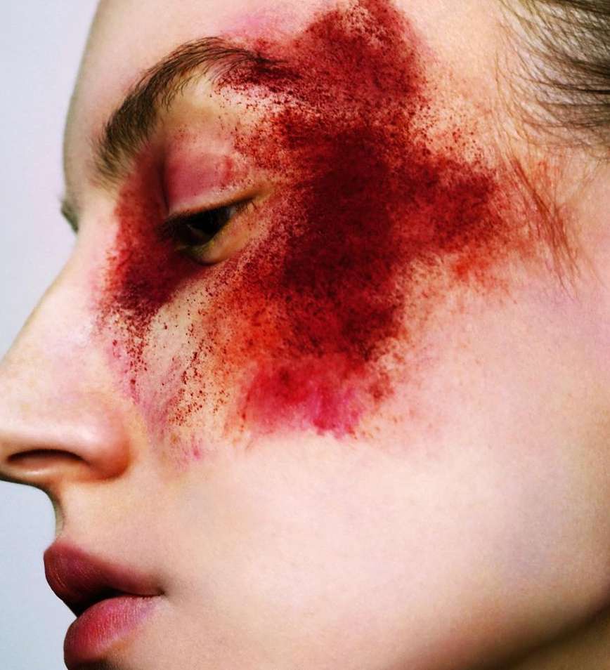 Close-up profile of a model's face with dramatic red pigment powder smeared across the eye and cheek.