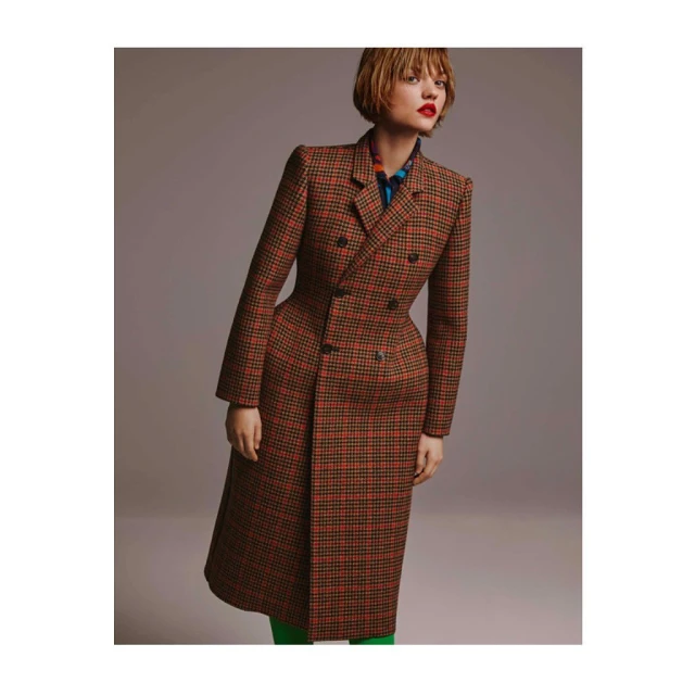 A young woman is posing in a studio. She is wearing a double-breasted plaid coat, a scarf, and green stockings. She has red lipstick.
