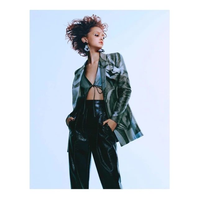 Fashion editorial shot of a woman in a blazer and shiny pants with dark hair blowing, against a light blue backdrop in a photo studio.