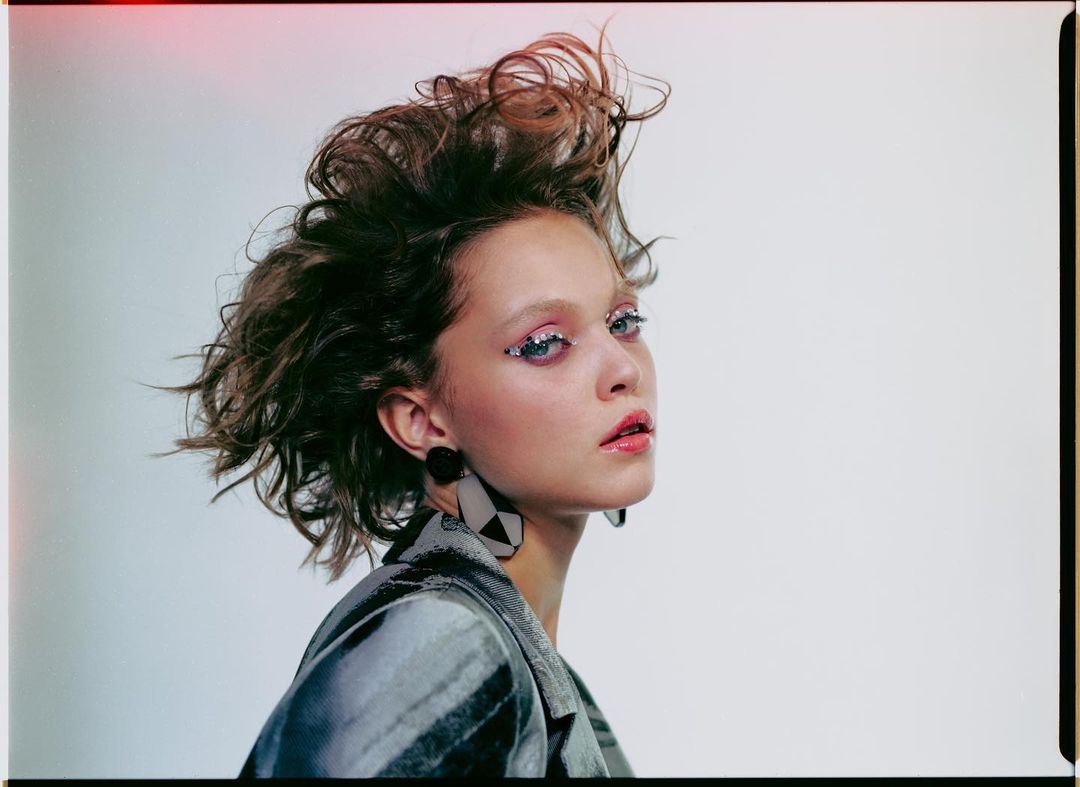 Portrait of a woman with styled hair, statement earrings, and a patterned jacket in a studio setting with a neutral background.