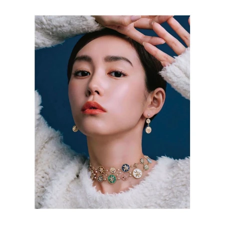 Adult woman poses indoors wearing a white sweater, decorative necklace, and matching earrings against a dark blue background.