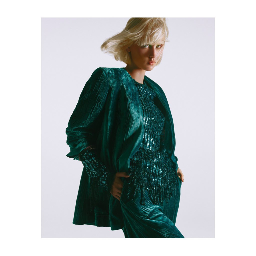 A woman with short blonde hair models a textured green velvet suit and matching top in a studio setting with soft, diffused lighting.