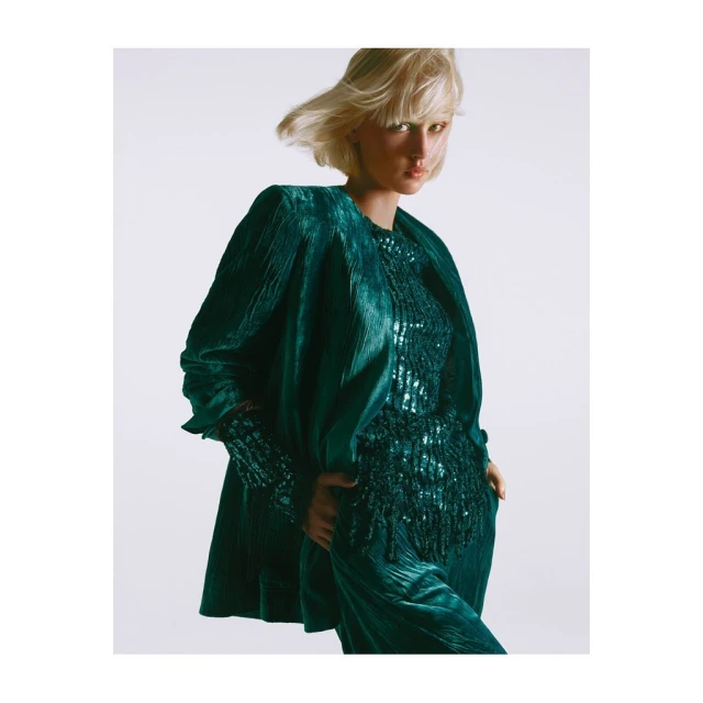 A woman with short blonde hair models a textured green velvet suit and matching top in a studio setting with soft, diffused lighting.