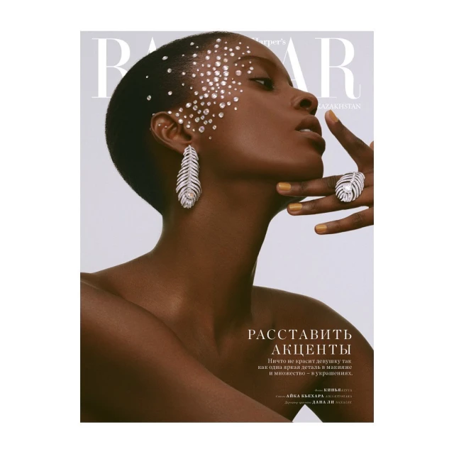 Close-up of a dark-skinned woman in profile wearing statement earrings and ring with crystals adhered to the face in a light studio.