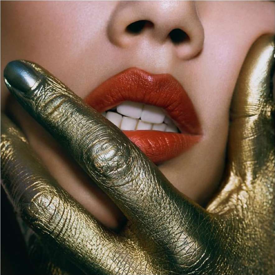 Close-up studio shot of an adult female face with red lipstick. A gold-painted hand covers part of the face in a Beauty-themed image.