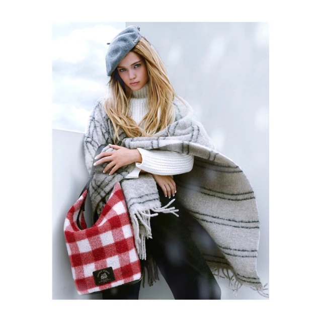 Adult female fashion model wearing a grey beret, white sweater, and grey striped shawl, holding a red and white checkered bag indoors.