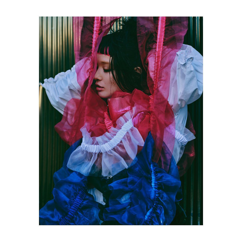Artistic studio portrait of a young woman with dark hair and light skin, draped in layers of white, red, and blue tulle fabric.