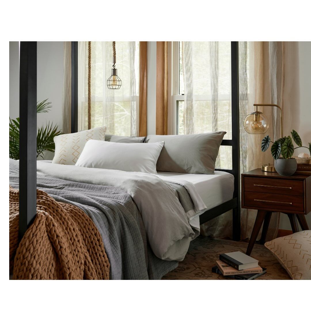 Bedroom interior with a four-poster bed, grey bedding, a brown knit throw, and a dark wood nightstand with a lamp and decor.
