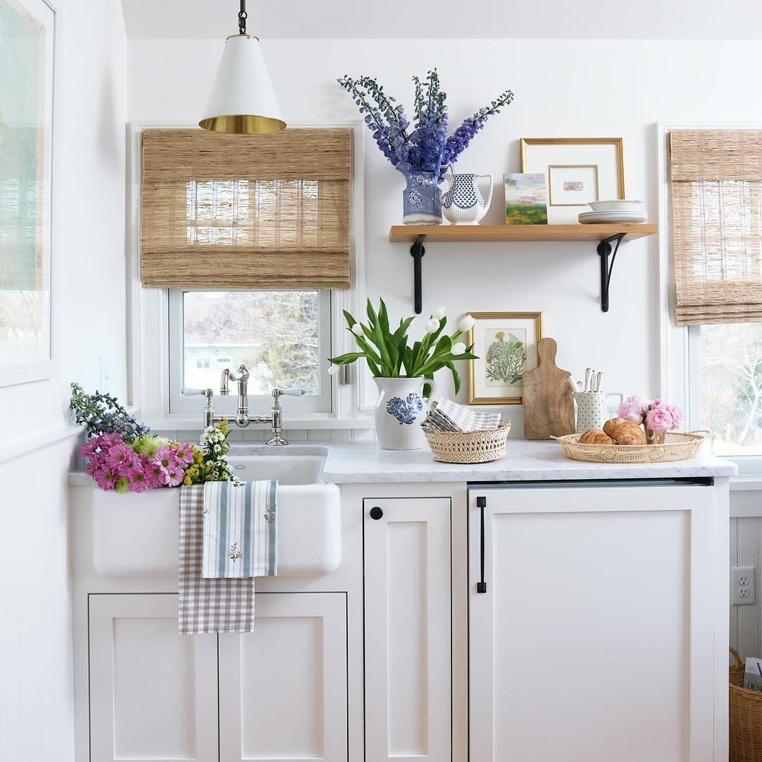 Bright kitchen with flowers, white cabinets, marble countertops, a farmhouse sink, and natural woven window shades.