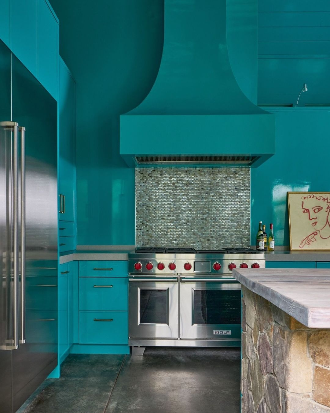Modern kitchen featuring turquoise cabinetry, stainless steel appliances, and a stone-clad kitchen island with a light countertop.