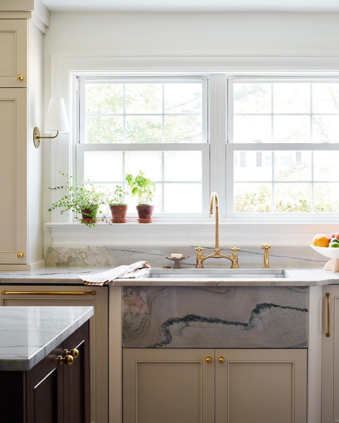 Kitchen interior featuring a marble farmhouse sink with gold faucet fixtures, positioned below a window with potted plants.