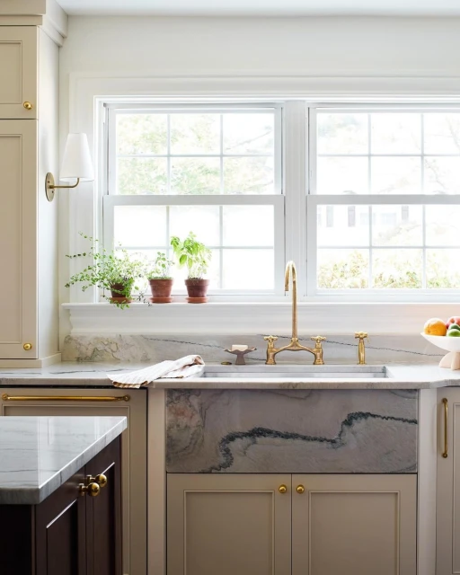 Kitchen interior featuring a marble farmhouse sink with gold faucet fixtures, positioned below a window with potted plants.