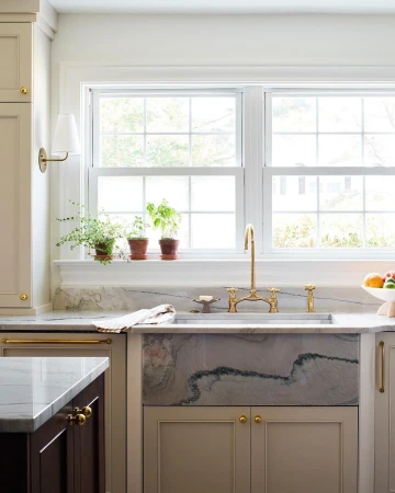 Kitchen interior featuring a marble farmhouse sink with gold faucet fixtures, positioned below a window with potted plants.